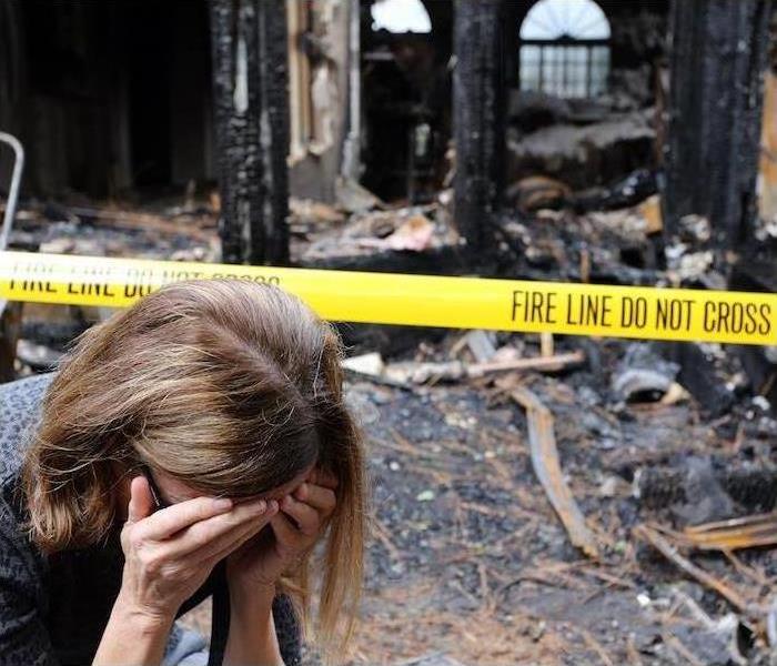 A woman holds her head in her hands in front of a burned-out house